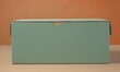© Nipon - A simple, rectangular green cardboard box with a small cutout on the top, placed on a light brown surface against a warm orange background.