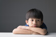 © bonnontawat - A young Asian boy with a sad expression, resting his head on folded arms, looking away against a plain background.