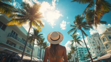 Wall Mural - A woman wearing a straw hat stands in front of a palm tree. She is wearing a bikini and is looking up at the sky