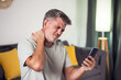 © Aleksej - Man with a beard sitting in a living room, touching his neck in discomfort, while holding a smartphone.