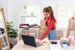 © Pichsakul - A young woman is holding several boxes while preparing orders for her online clothing business. She is smiling and working in a bright, organized workspace.