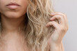 © Марина Демешко - A young blonde woman touching her wavy tangled unruly dry bleached hair on a light grey background. Hair care