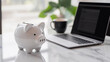 © Maksym - A minimalist white piggy bank on a clean, modern desk with a laptop and a cup of coffee, symbolizing professional financial planning and digital investment.