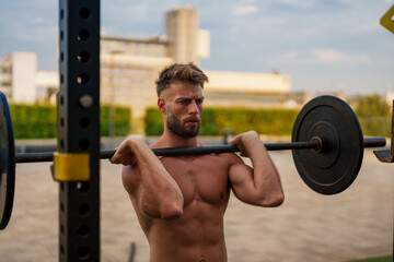 Handsome man lifting a barbell overhead during an outdoor workout. He is shirtless and appears focused on his exercise. The background shows an urban setting with buildings and greenery.