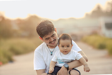 a father holding his baby boy faces the camera with a smile. father and baby boy look happy and enjoy the afternoon atmosphere.