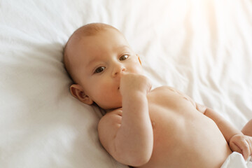 A peaceful infant lies comfortably on a white bed, sucking on their thumb while enjoying the warmth of sunlight filtering through the window.