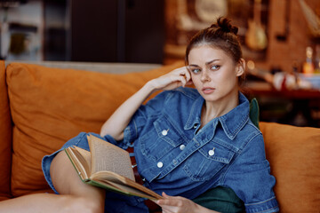 Young woman in denim dress sitting on couch, engrossed in book, gazes confidently at camera