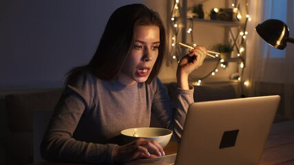 Poster - A woman sits at a table, eating from a bowl and focused on her laptop in a dimly lit room with warm decorations.