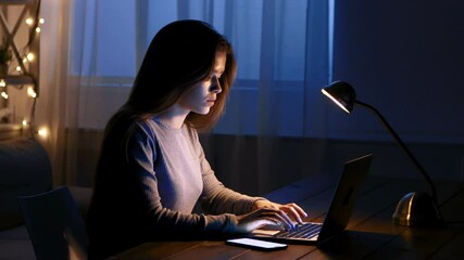 Poster - A woman focuses on her laptop while seated at a desk, illuminated by a desk lamp in a cozy, dark room.