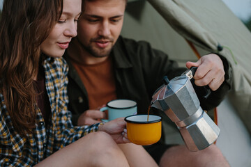 Wall Mural - Smiling young man and woman drinking delicious coffee sitting near their tent at sunset. The concept of recreation outside the city.