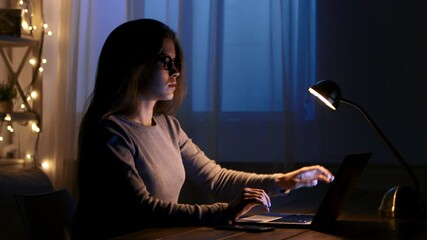 Poster - A woman sits at a desk working on her laptop in a dark room illuminated by a desk lamp.