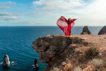 Wall Mural - A woman in a red dress stands on a rocky cliff overlooking the ocean. The scene is serene and peaceful, with the woman's flowing dress adding a sense of grace and beauty to the landscape.