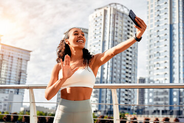 Happy smiling cheerful female fitness trainer in sportswear holding phone and greeting followers, friends online while outside after morning run. Social networks, communication, sports lifestyle..