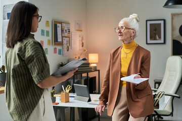 Poster - Elderly lady engaging in conversation with younger woman in contemporary office environment holding papers while standing beside desk and notes on wall