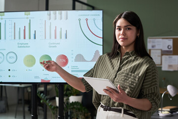 Poster - Woman pointing at a presentation with business metrics displayed on screen, holding a tablet and showcasing various growth indicators in office setting