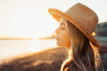 Wall Mural - Portrait of one young woman wearing hat at the beach closing eyes enjoying free time and freedom outdoors. Having fun relaxing and living happy moments.