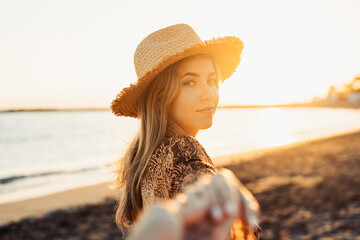 Wall Mural - One cute girl walking on the sand of the beach holding her boyfriend’s hand. Enjoying travel and vacation summer lifestyle together outdoors at sunset.