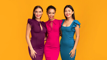 It Girls. Three Diverse Young Ladies In Cocktail Dresses Smiling At Camera And Hugging Standing On Yellow Background. Studio Shot