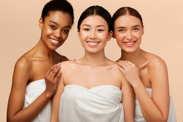 Wall Mural - Beauty And Skin Care. Three Diverse Women In Bath Towels Smiling At Camera Posing Over Beige Background. Studio Shot