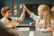 © Seventyfour - Side view of young blonde schoolgirl sitting at desk happily giving high five to male classmate while having fun with friends during break at school