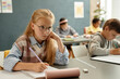 © Seventyfour - Medium shot of studious young girl with glasses sitting at desk attentively looking at blackboard while writing in exercise book in classroom with blue wall, copy space