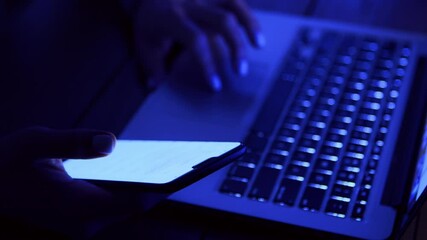 Poster - A person types on a laptop with one hand while holding a smartphone in the other, illuminated by a faint blue light.