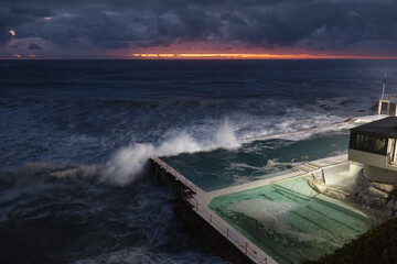 This photo captures a dramatic seascape at dusk with waves crashing over a seaside pool, juxtaposed against a tranquil sky with hints of sunset. The scene blends natural beauty and human-made structur