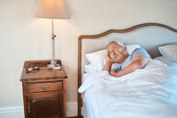 Senior man, sleeping and bed with lamp light for morning, rest or recovery in retirement home. Elderly, male person or asleep with blankets, sheets or duvet by pedestal for relaxation in bedroom