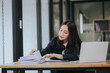 © Witoon - A cheerful young Asian businesswoman sits at her modern desk, typing on a laptop, working on corporate projects with paperwork, smiling in a busy workplace.