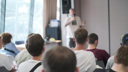 Wall Mural - Group of people sitting and listening to a speaker during a business presentation. The setting is a bright, modern conference room with large windows.