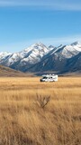 Fototapeta Góry - A white RV with solar panels rests in golden grassland, surrounded by majestic snow-capped mountains and under a dramatic cloudy sky, perfect for outdoor living.