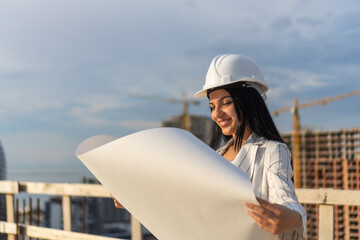 Portrait of a smiling young beautiful woman architect, design engineer, manager in a hard hat with drawings on whatman paper in her hands on a high floor of a construction site, copy space.