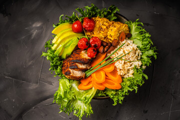 Buddha bowl with vegetables, healthy salad with avocado, carrots, corn and rice, baked chicken breast on a black background with a burgundy napkin and fork. Healthy eating concept. Copyspace. High