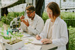 © AnnaStills - Two scientists working in a lab examining plant specimens surrounded by lush greenery. Researching plant growth under controlled conditions in a greenhouse setting