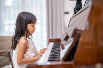 Young asian girl learning to play piano at home, practicing music on a classic upright piano developing musical skills, childhood education indoor lifestyle, asian kid with long hair in casual dress