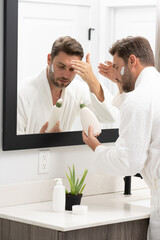 A man is applying lotion to his face in front of a mirror. Concept of self-care and personal grooming