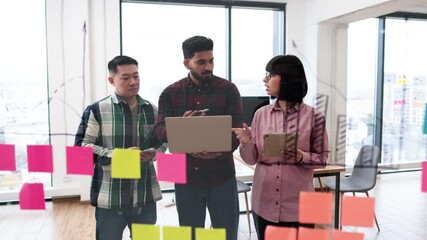 Wall Mural - Team collaboration with sticky notes and laptop in modern office environment. Group of professionals brainstorming, discussing ideas, and planning project on glass board with colorful notes.