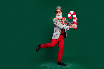 Sticker - Full body portrait of excited aged man blazer decorated xmas tree toys red pants cane stick candy isolated on dark green color background