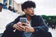 © BullRun - Contemplative male tourist in casual apparel resting at city urban setting pondering on content ideas for publication in social networks, millennial pensive Latino teenager with mobile phone