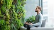 © dakinya - A man practicing meditation in front of a lush green vertical garden inside a modern eco-friendly office, improving mental health and focus, surrounded by natural light, selective focus