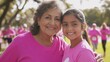 © suyu - Senior Hispanic woman smiling while hugging her young adult granddaughter at a breast cancer awareness charity race, both wearing pink athletic clothing as other participants register in background