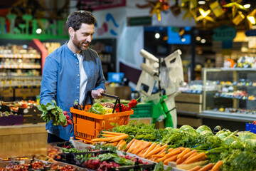 Man browsing fresh vegetables in grocery store holding orange shopping basket filled with assorted produce including lettuce, radishes, and peppers.