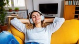 A young woman sits back on a mustard-colored sofa, eyes closed and completely immersed in music from her headphones, radiating joy and relaxation.