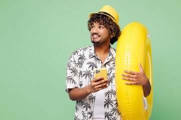 Sticker - Young happy Indian man wear beach shirt hat rest near hotel pool hold yellow rubber ring use mobile cell phone look aside isolated on plain green color background. Summer vacation sea sun tan concept.