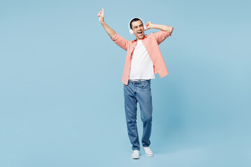 Wall Mural - Full body young middle eastern man he wearing pink shirt white t-shirt listen to music in headphones raise up hand dance isolated on plain pastel light blue cyan background studio. Lifestyle concept.