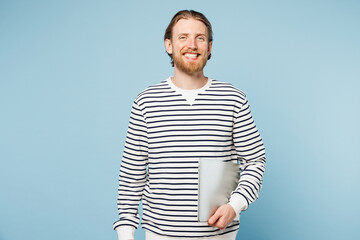 Sticker - Young smiling fun cheerful happy IT man he wear striped sweatshirt casual clothes hold closed laptop pc computer isolated on plain pastel light blue cyan background studio portrait. Lifestyle concept.