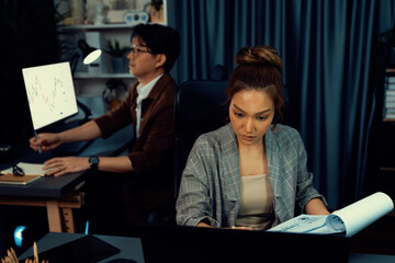 Businesswoman with stressful face looking document paperwork file while coworker trading stock market at back at over late night time at modern office, working on startup company project. Infobahn.