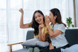 © amnaj - Asian mother and daughter sitting on a sofa, sharing an exciting secret and raising their fists in the air