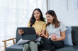 © amnaj - Two asian women happily sharing on a tablet in a cozy home, bonding and enjoying quality time together in a modern living room filled with natural light