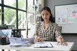 © amnaj - Young professional analyzes financial data at her desk, with paperwork and a whiteboard showing business metrics